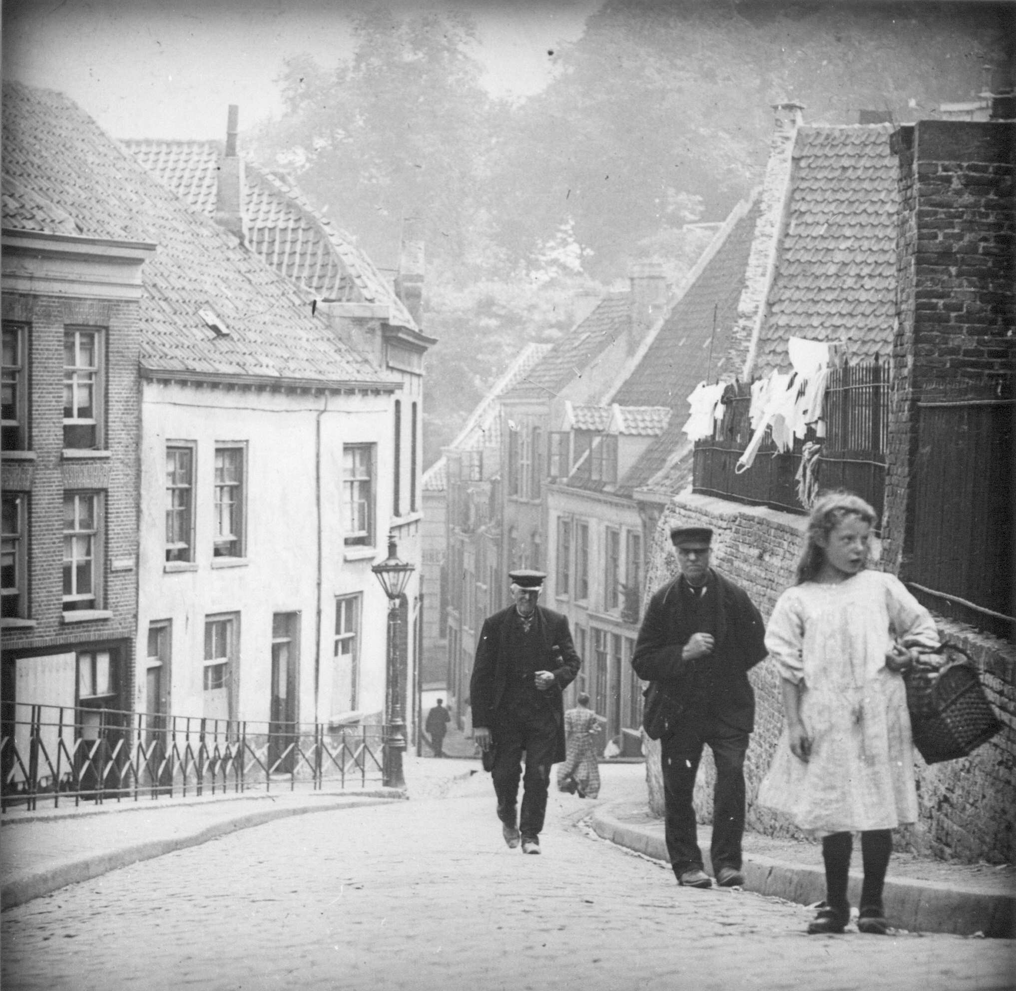 De Vleeshouderstraat rond 1900. Op de foto een meisje in een wit jurkje met een mand aan haar linker arm. Op de achtergrond 2 oudere mannen met een pet.